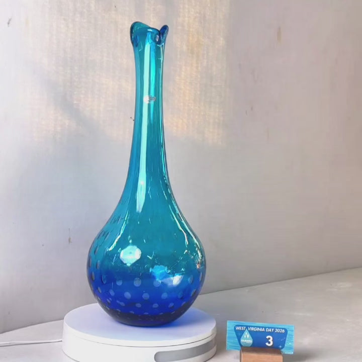 A translucent blue glass vase with a flared neck and dotted pattern, displayed on a white circular base against a light gray wall.