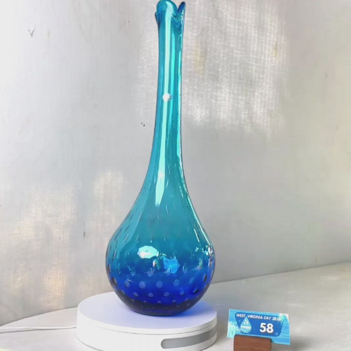 A translucent blue glass vase with a flared neck and dotted pattern, displayed on a white circular base against a light gray wall.