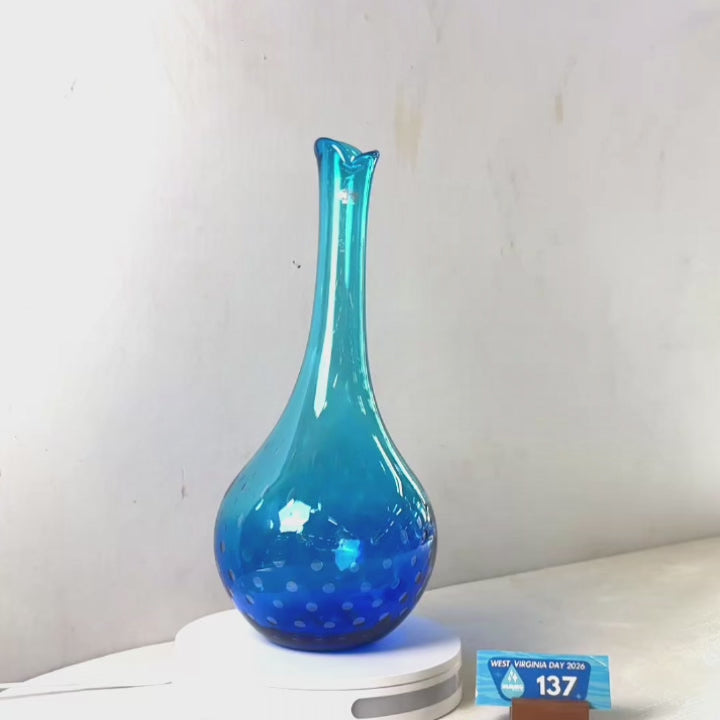 A translucent blue glass vase with a flared rim and dotted pattern near the base, displayed on a white circular stand against a plain light gray wall.