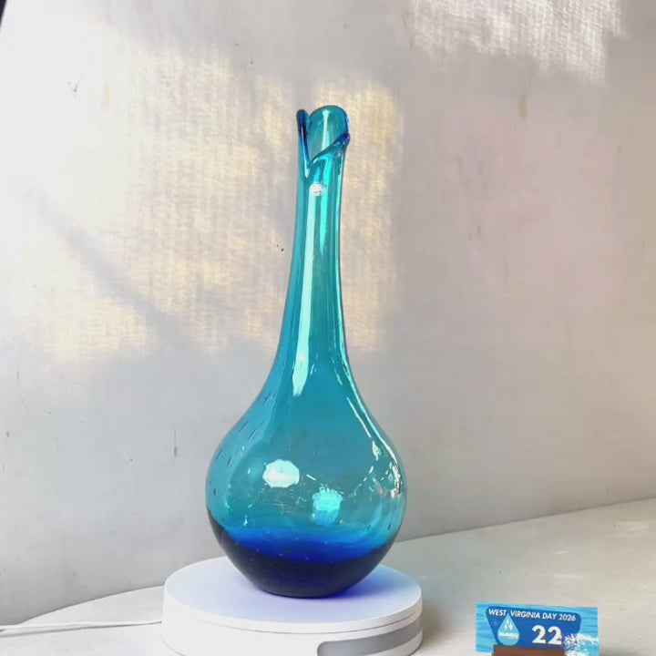 A translucent blue glass vase with a flared neck and dotted pattern, displayed on a white circular base against a light gray wall.