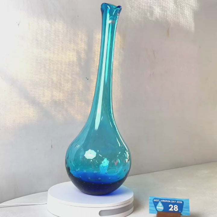 A translucent blue glass vase with a flared neck and dotted pattern, displayed on a white circular base against a light gray wall.