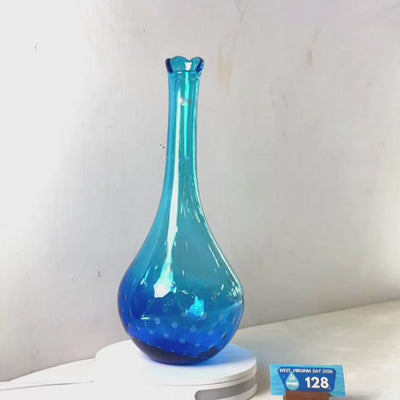 A translucent blue glass vase with a flared neck and dotted pattern near the base, displayed on a white circular light box. A small wrapped item and a brown block are placed beside it on a white surface against a plain light gray wall.