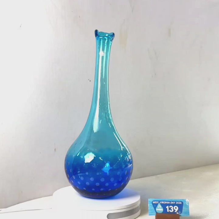 A translucent blue glass vase with a flared rim and dotted pattern near the base, displayed on a white circular stand against a plain light gray wall.