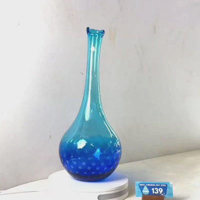 A translucent blue glass vase with a flared rim and dotted pattern near the base, displayed on a white circular stand against a plain light gray wall.