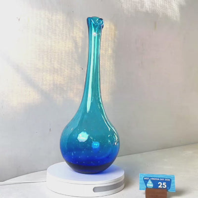 A translucent blue glass vase with a flared neck and dotted pattern, displayed on a white circular base against a light gray wall.
