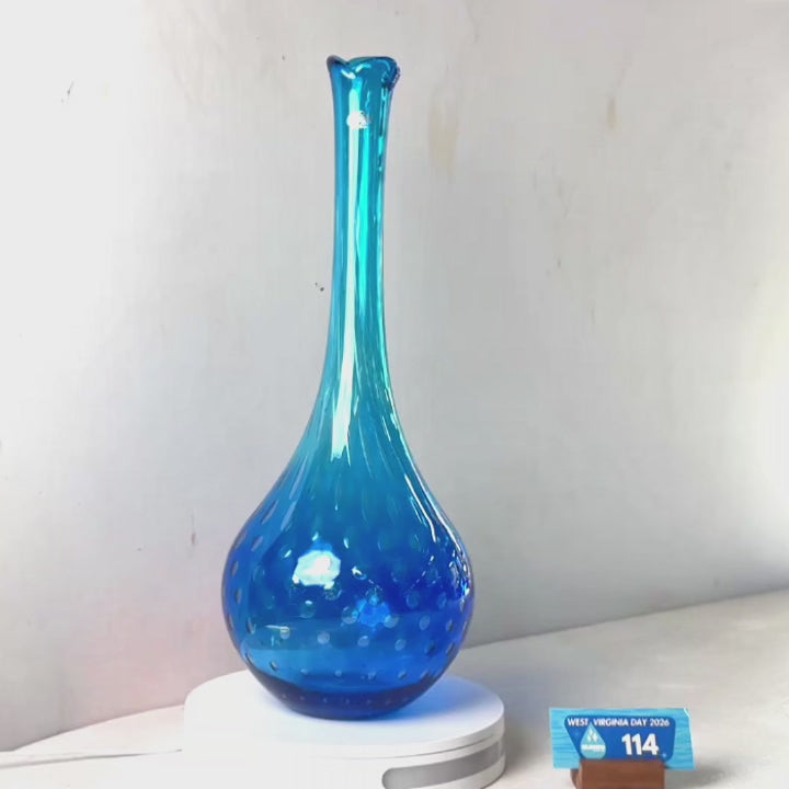 A translucent blue glass vase with a flared neck and dotted pattern near the base, displayed on a white circular light box. A small wrapped item and a brown block are placed beside it on a white surface against a plain light gray wall.