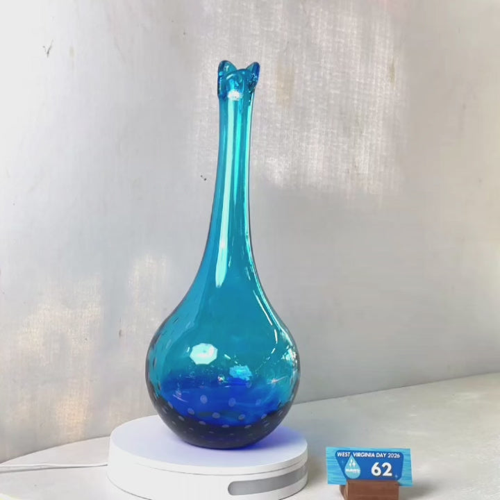 A translucent blue glass vase with a flared neck and dotted pattern, displayed on a white circular base against a light gray wall.