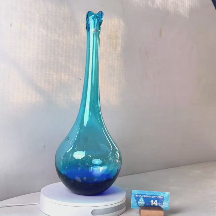 A translucent blue glass vase with a flared neck and dotted pattern, displayed on a white circular base against a light gray wall.