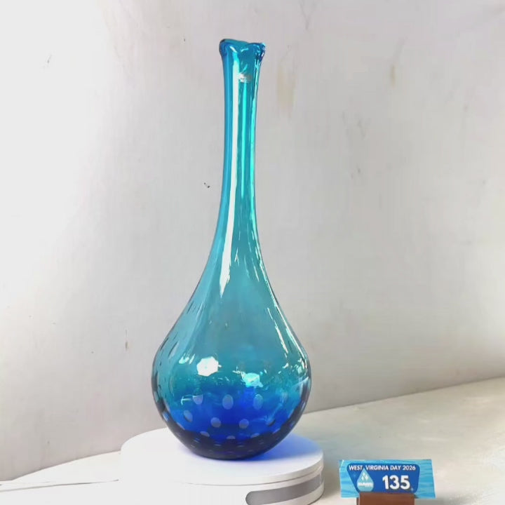 A translucent blue glass vase with a flared rim and dotted pattern near the base, displayed on a white circular stand against a plain light gray wall.