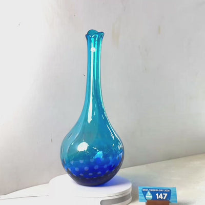 A translucent blue glass vase with a flared rim and dotted pattern near the base, displayed on a white circular stand against a plain light gray wall.