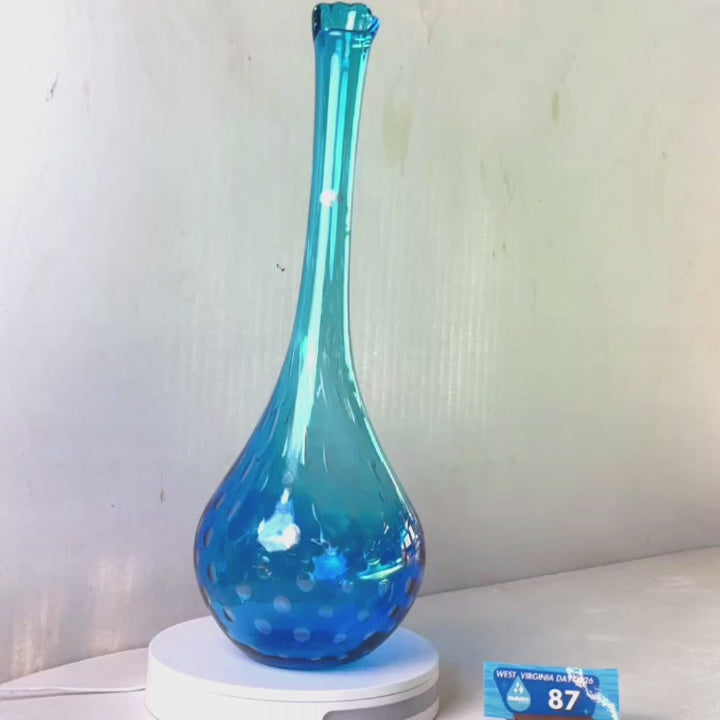A translucent blue glass vase with a flared rim and subtle white speckled pattern near the base, displayed on a white round scale with a small price tag in front, against a plain light gray wall.
