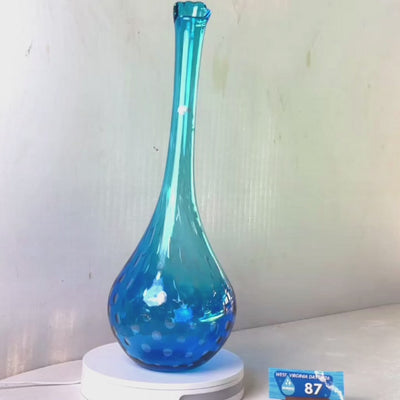 A translucent blue glass vase with a flared rim and subtle white speckled pattern near the base, displayed on a white round scale with a small price tag in front, against a plain light gray wall.