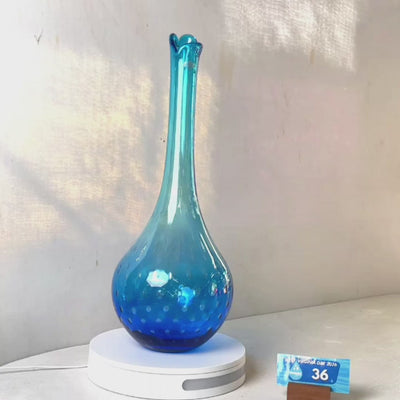 A translucent blue glass vase with a flared neck and dotted pattern, displayed on a white circular base against a light gray wall.