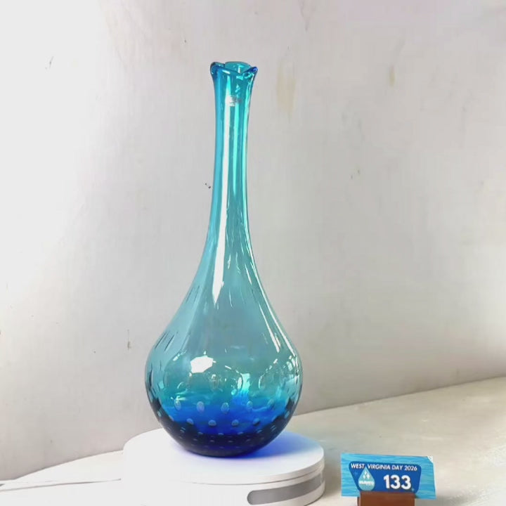 A translucent blue glass vase with a flared rim and dotted pattern near the base, displayed on a white circular stand against a plain light gray wall.