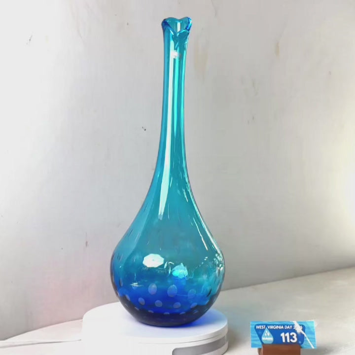 A translucent blue glass vase with a flared neck and dotted pattern near the base, displayed on a white circular light box. A small wrapped item and a brown block are placed beside it on a white surface against a plain light gray wall.