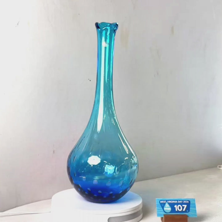 A translucent blue glass vase with a flared rim and dotted pattern near the base, displayed on a white circular stand against a plain light gray wall.