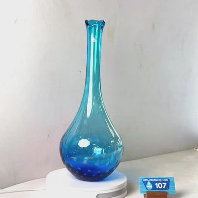 A translucent blue glass vase with a flared rim and dotted pattern near the base, displayed on a white circular stand against a plain light gray wall.