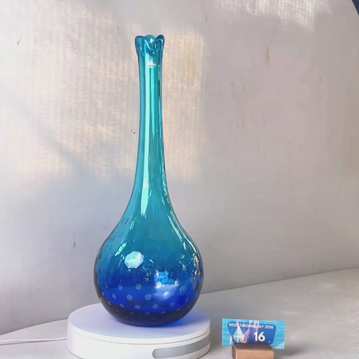 A translucent blue glass vase with a flared neck and dotted pattern, displayed on a white circular base against a light gray wall.