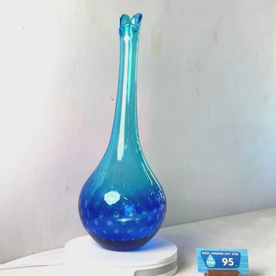 A translucent blue glass vase with a flared rim and subtle white speckled pattern near the base, displayed on a white round scale with a small price tag in front, against a plain light gray wall.