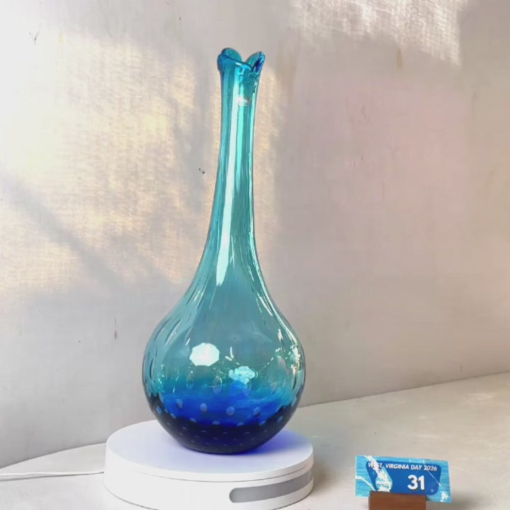 A translucent blue glass vase with a flared neck and dotted pattern, displayed on a white circular base against a light gray wall.