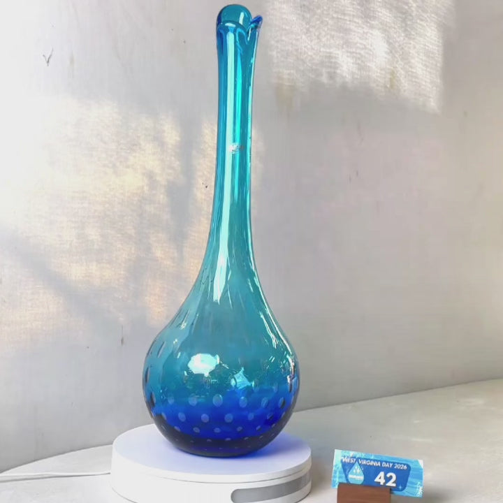 A translucent blue glass vase with a flared neck and dotted pattern, displayed on a white circular base against a light gray wall.
