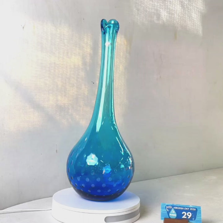 A translucent blue glass vase with a flared neck and dotted pattern, displayed on a white circular base against a light gray wall.