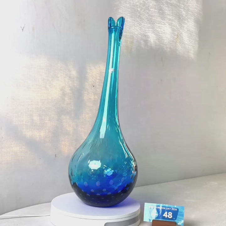 A translucent blue glass vase with a flared neck and dotted pattern, displayed on a white circular base against a light gray wall.