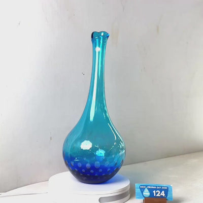 A translucent blue glass vase with a flared neck and dotted pattern near the base, displayed on a white circular light box. A small wrapped item and a brown block are placed beside it on a white surface against a plain light gray wall.