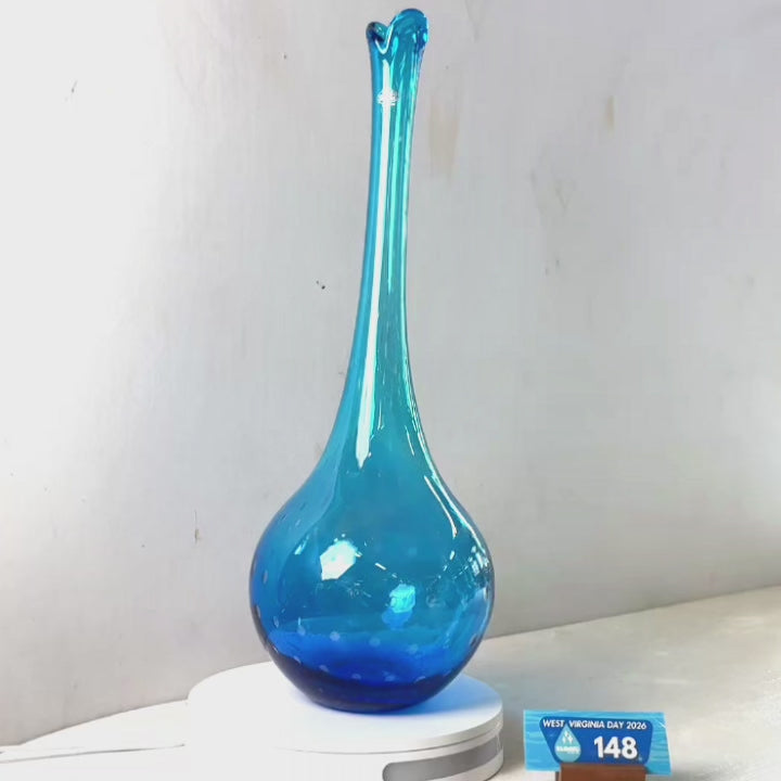 A translucent blue glass vase with a flared rim and dotted pattern near the base, displayed on a white circular stand against a plain light gray wall.
