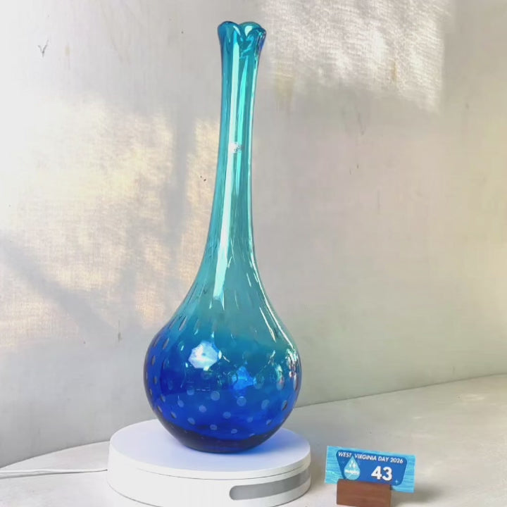 A translucent blue glass vase with a flared neck and dotted pattern, displayed on a white circular base against a light gray wall.