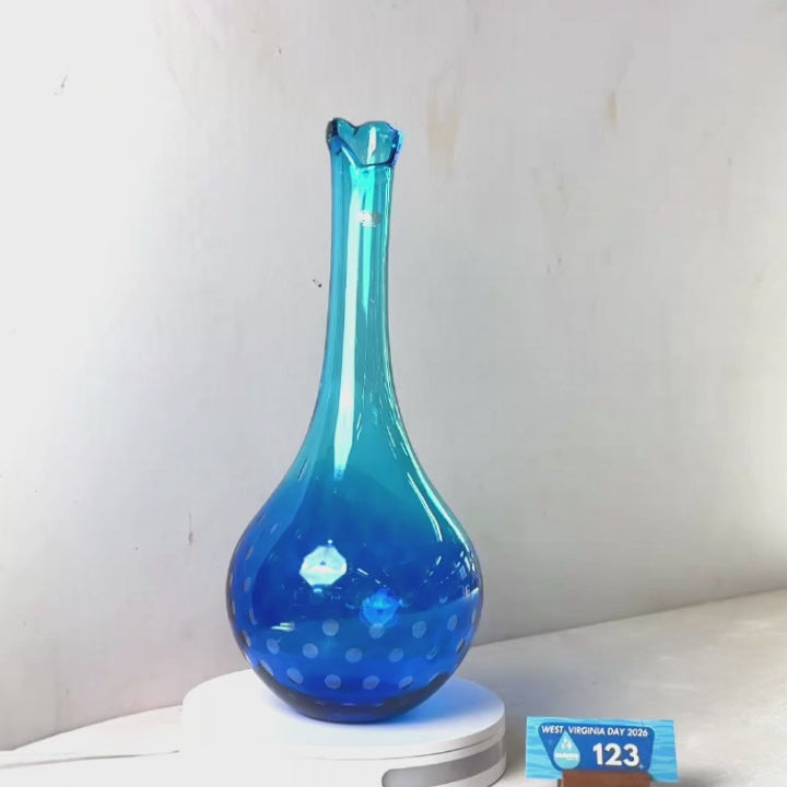 A translucent blue glass vase with a flared neck and dotted pattern near the base, displayed on a white circular light box. A small wrapped item and a brown block are placed beside it on a white surface against a plain light gray wall.
