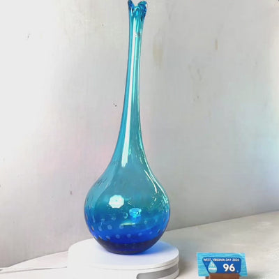 A translucent blue glass vase with a flared rim and subtle white speckled pattern near the base, displayed on a white round scale with a small price tag in front, against a plain light gray wall.