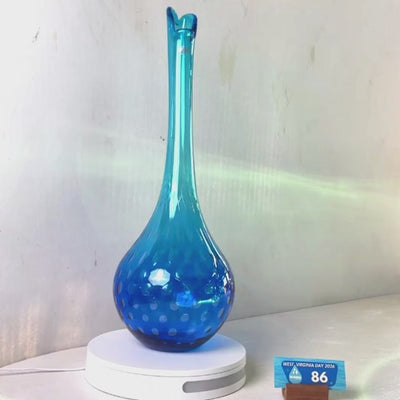 A translucent blue glass vase with a flared rim and subtle white speckled pattern near the base, displayed on a white round scale with a small price tag in front, against a plain light gray wall.