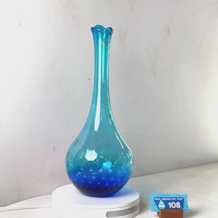 A translucent blue glass vase with a flared rim and dotted pattern near the base, displayed on a white circular stand against a plain light gray wall.