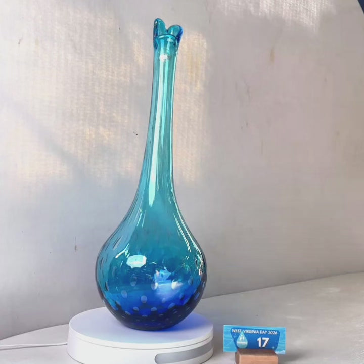 A translucent blue glass vase with a flared neck and dotted pattern, displayed on a white circular base against a light gray wall.