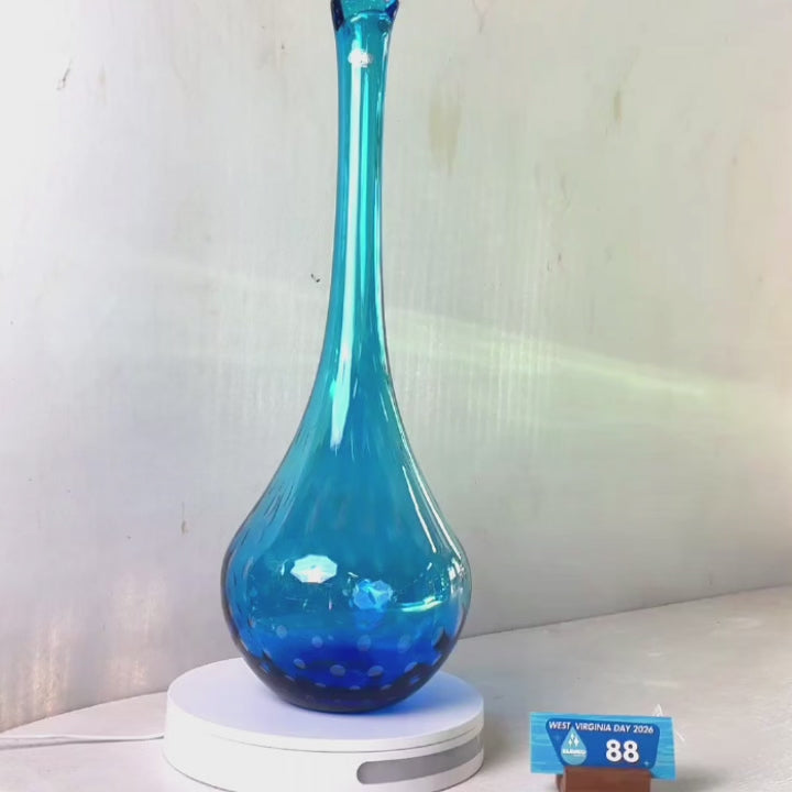 A translucent blue glass vase with a flared rim and subtle white speckled pattern near the base, displayed on a white round scale with a small price tag in front, against a plain light gray wall.