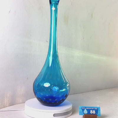 A translucent blue glass vase with a flared rim and subtle white speckled pattern near the base, displayed on a white round scale with a small price tag in front, against a plain light gray wall.