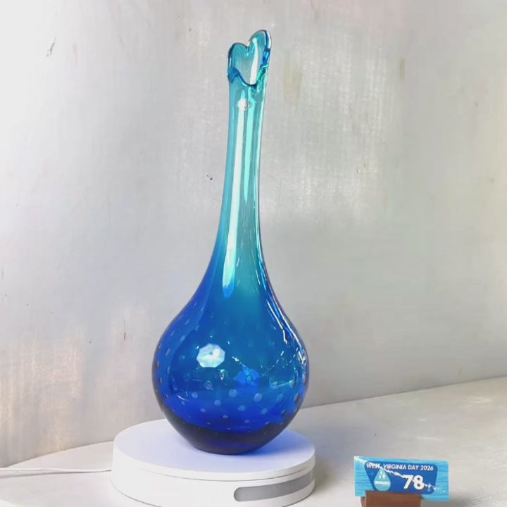A translucent blue glass vase with a flared rim and subtle white speckled pattern near the base, displayed on a white round scale with a small price tag in front, against a plain light gray wall.