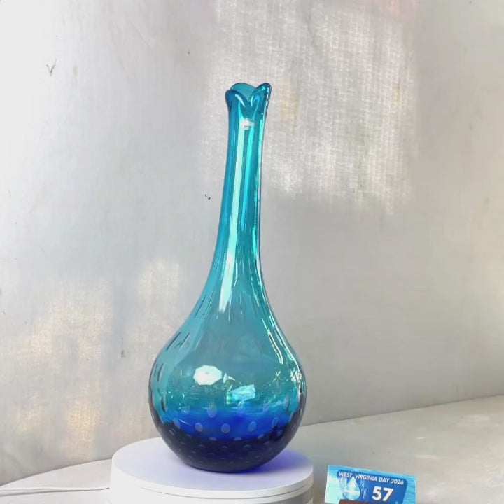 A translucent blue glass vase with a flared neck and dotted pattern, displayed on a white circular base against a light gray wall.