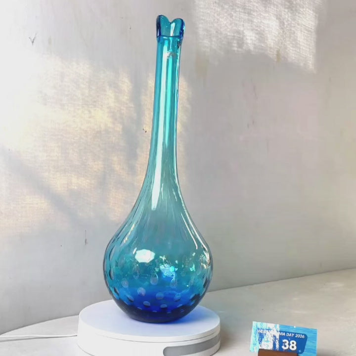 A translucent blue glass vase with a flared neck and dotted pattern, displayed on a white circular base against a light gray wall.
