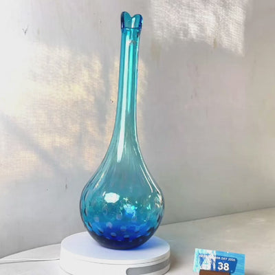 A translucent blue glass vase with a flared neck and dotted pattern, displayed on a white circular base against a light gray wall.