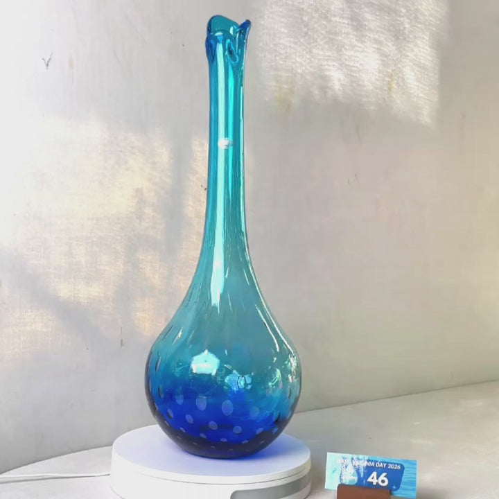 A translucent blue glass vase with a flared neck and dotted pattern, displayed on a white circular base against a light gray wall.