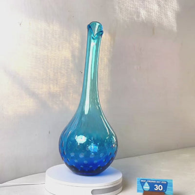 A translucent blue glass vase with a flared neck and dotted pattern, displayed on a white circular base against a light gray wall.