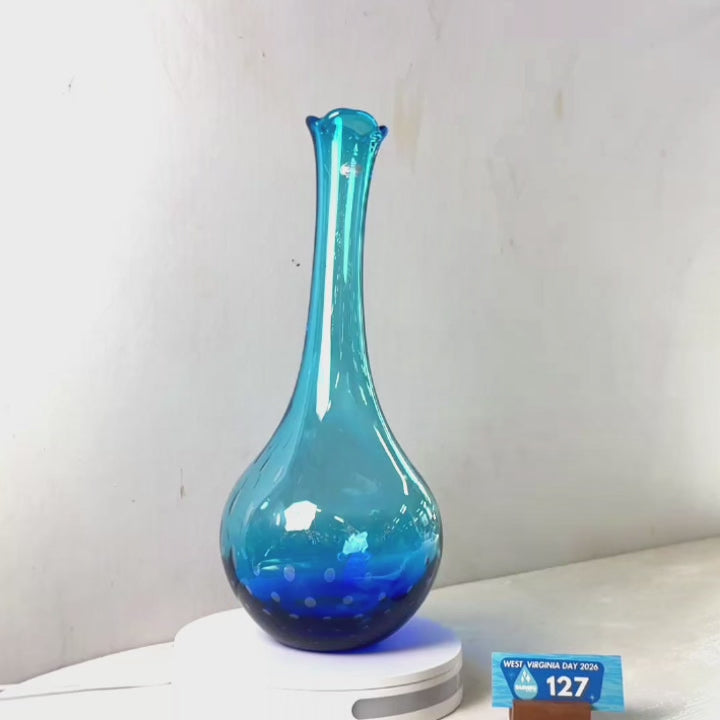 A translucent blue glass vase with a flared neck and dotted pattern near the base, displayed on a white circular light box. A small wrapped item and a brown block are placed beside it on a white surface against a plain light gray wall.