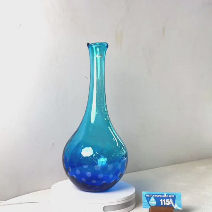 A translucent blue glass vase with a flared neck and dotted pattern near the base, displayed on a white circular light box. A small wrapped item and a brown block are placed beside it on a white surface against a plain light gray wall.