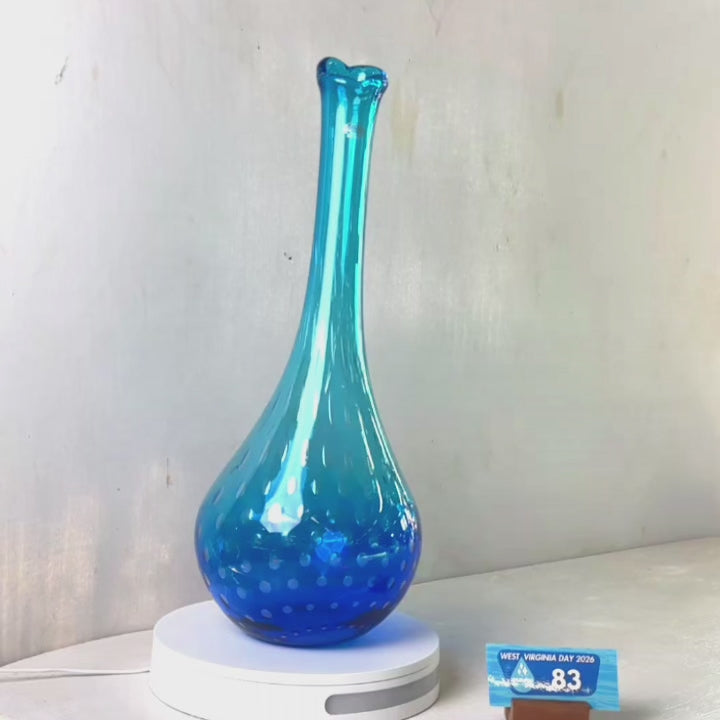 A translucent blue glass vase with a flared rim and subtle white speckled pattern near the base, displayed on a white round scale with a small price tag in front, against a plain light gray wall.