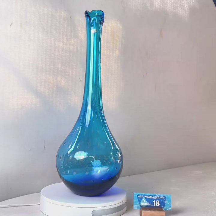 A translucent blue glass vase with a flared neck and dotted pattern, displayed on a white circular base against a light gray wall.