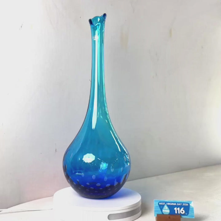 A translucent blue glass vase with a flared neck and dotted pattern near the base, displayed on a white circular light box. A small wrapped item and a brown block are placed beside it on a white surface against a plain light gray wall.