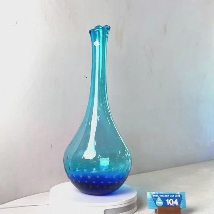 A translucent blue glass vase with a flared rim and dotted pattern near the base, displayed on a white circular stand against a plain light gray wall.