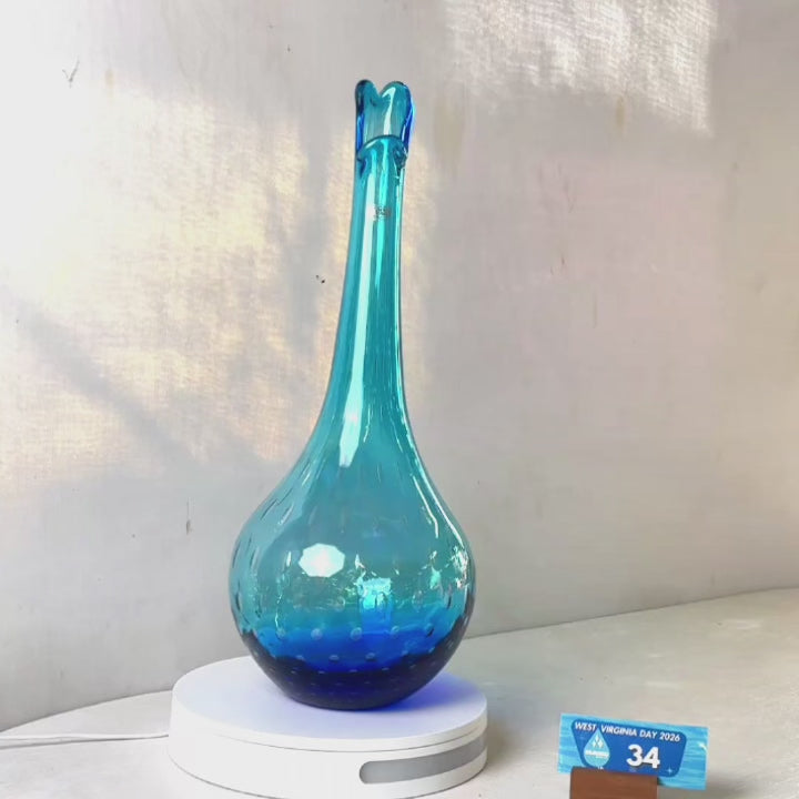 A translucent blue glass vase with a flared neck and dotted pattern, displayed on a white circular base against a light gray wall.
